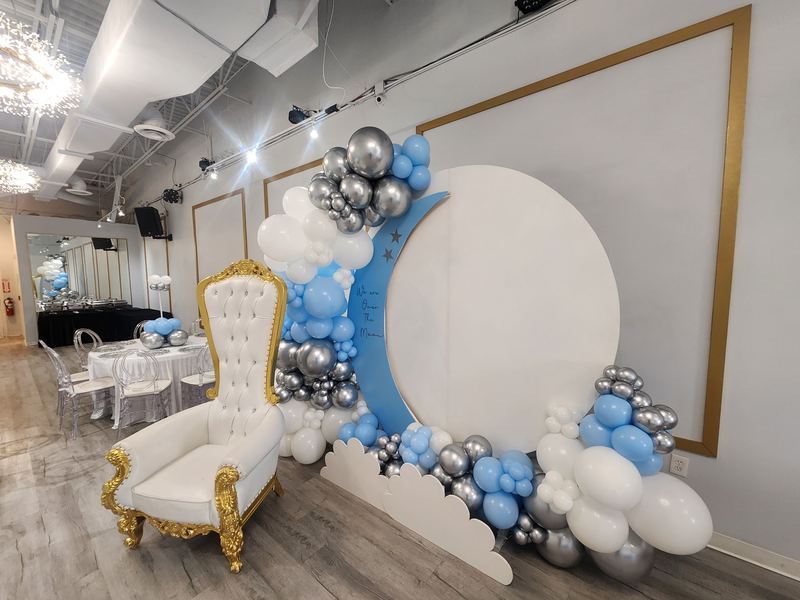 Baby shower backdrop with a blue moon, “We are over the moon” sign, blue, white and silver balloons and a white and gold chair