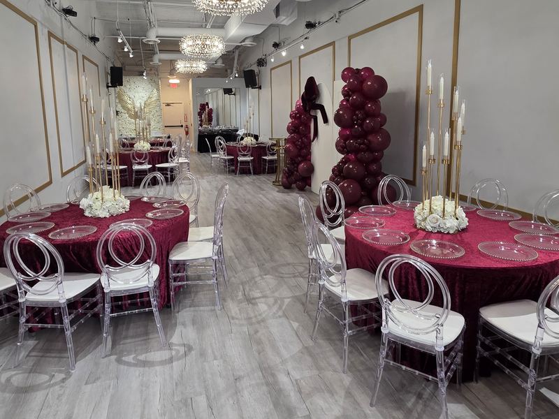 Mesas redondas con manteles de terciopelo color vino, candelabros dorados, bajoplatos transparentes y una torre de globos color vino