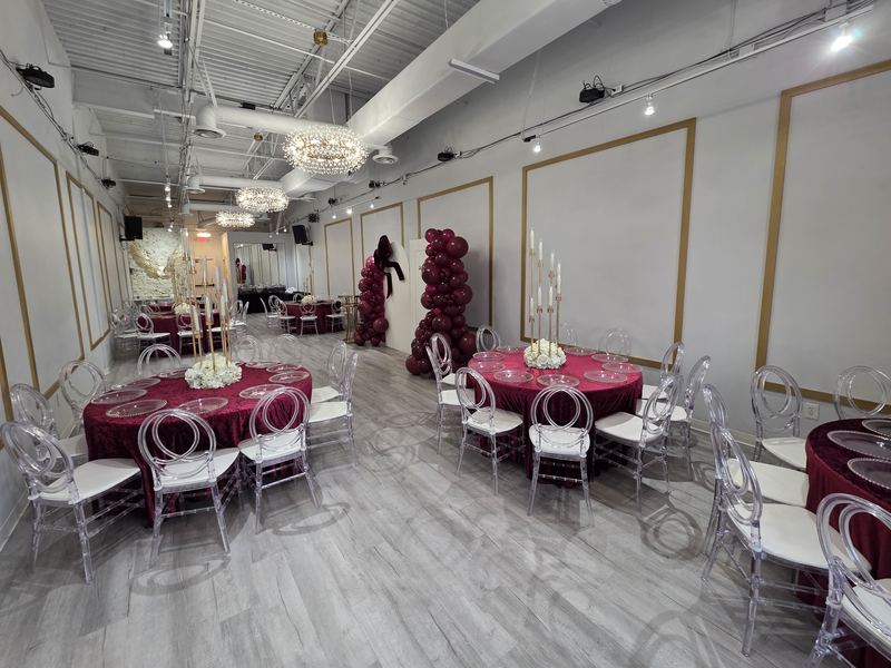 Vista del salón completo: mesas redondas con manteles de terciopelo color vino, candelabros dorados altos con flores blancas y torres de globos color vino