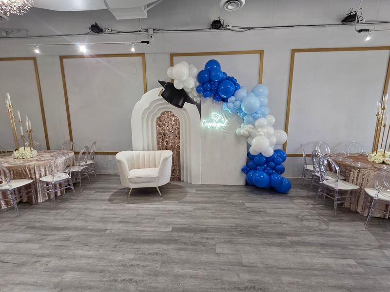 Fondo de graduación con paneles blancos en arco, letrero de neón y globos azul rey, blancos y negros junto a un sillón blanco