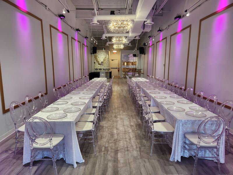 Mesas largas tipo banquete con manteles blancos y sillas transparentes bajo luces moradas
