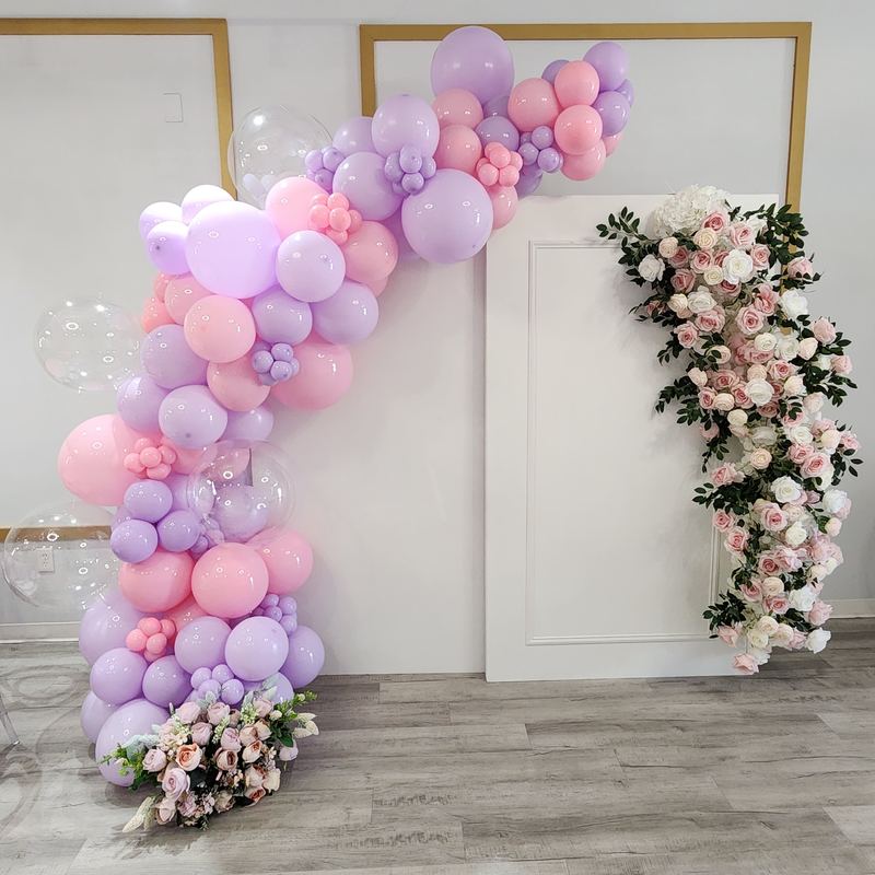 Guirnalda de globos rosas y lilas con una columna de rosas rosas y blancas sobre paneles blancos en arco