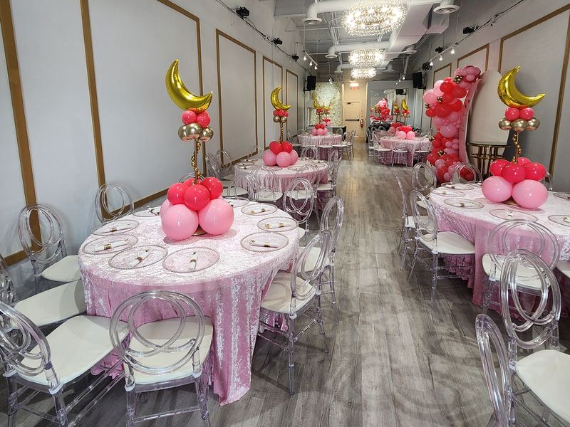 Vista del salón completo: mesas redondas con manteles rosas de terciopelo y centros de mesa de globos rosas con lunas doradas