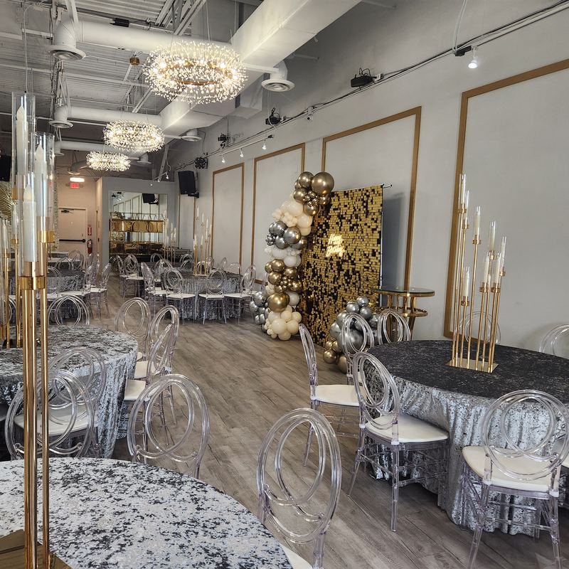 El salón completo con mesas plateadas, candelabros dorados altos y lámparas de cristal, con una pared de lentejuelas doradas y globos al fondo