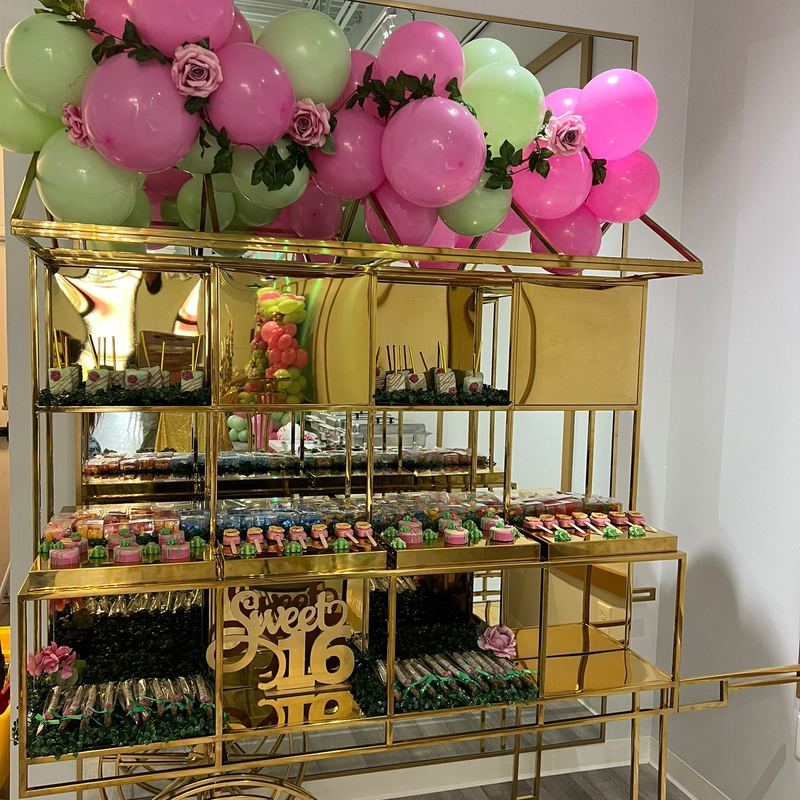 Carrito dorado de postres con letras doradas Sweet 16 bajo globos rosas y verdes