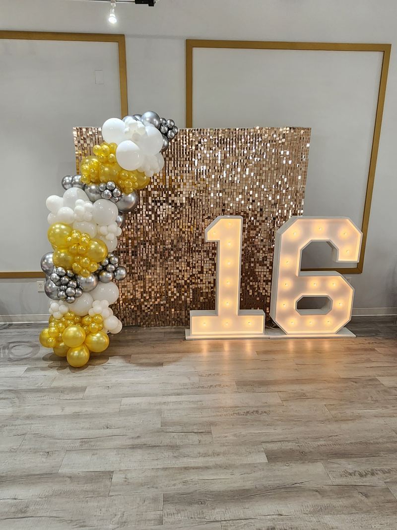 Número 16 luminoso frente a una pared de lentejuelas doradas con globos dorados, blancos y plateados