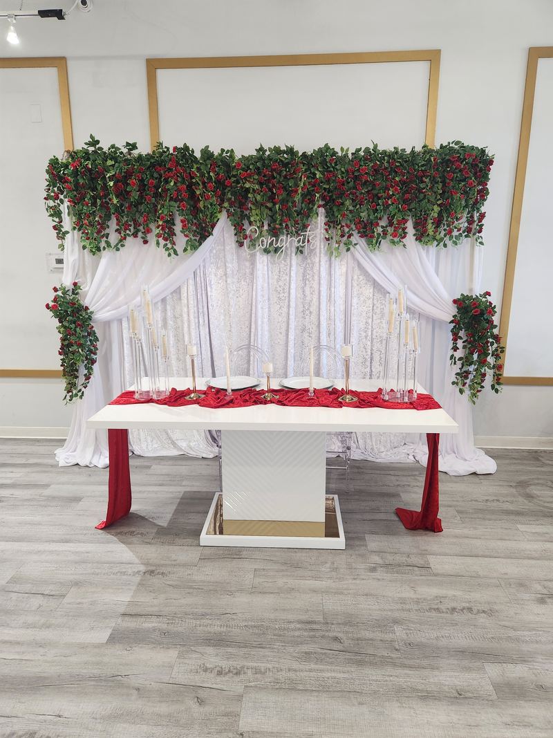 Head table draped in white with a red runner and a garland of red roses and greenery